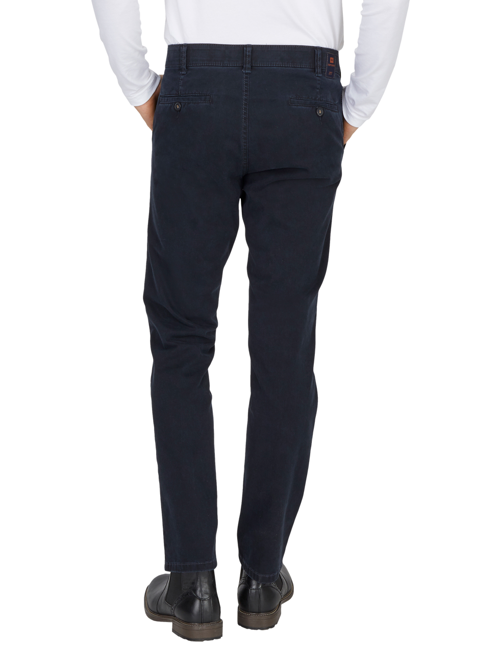 Blick auf einen Mann, der dunkelblaue, schmale Chino-Hose trägt, die hinten sichtbar ist. Er hat die Hände in den Taschen, trägt eine weiße Hemdjacke und schwarze Schuhe.