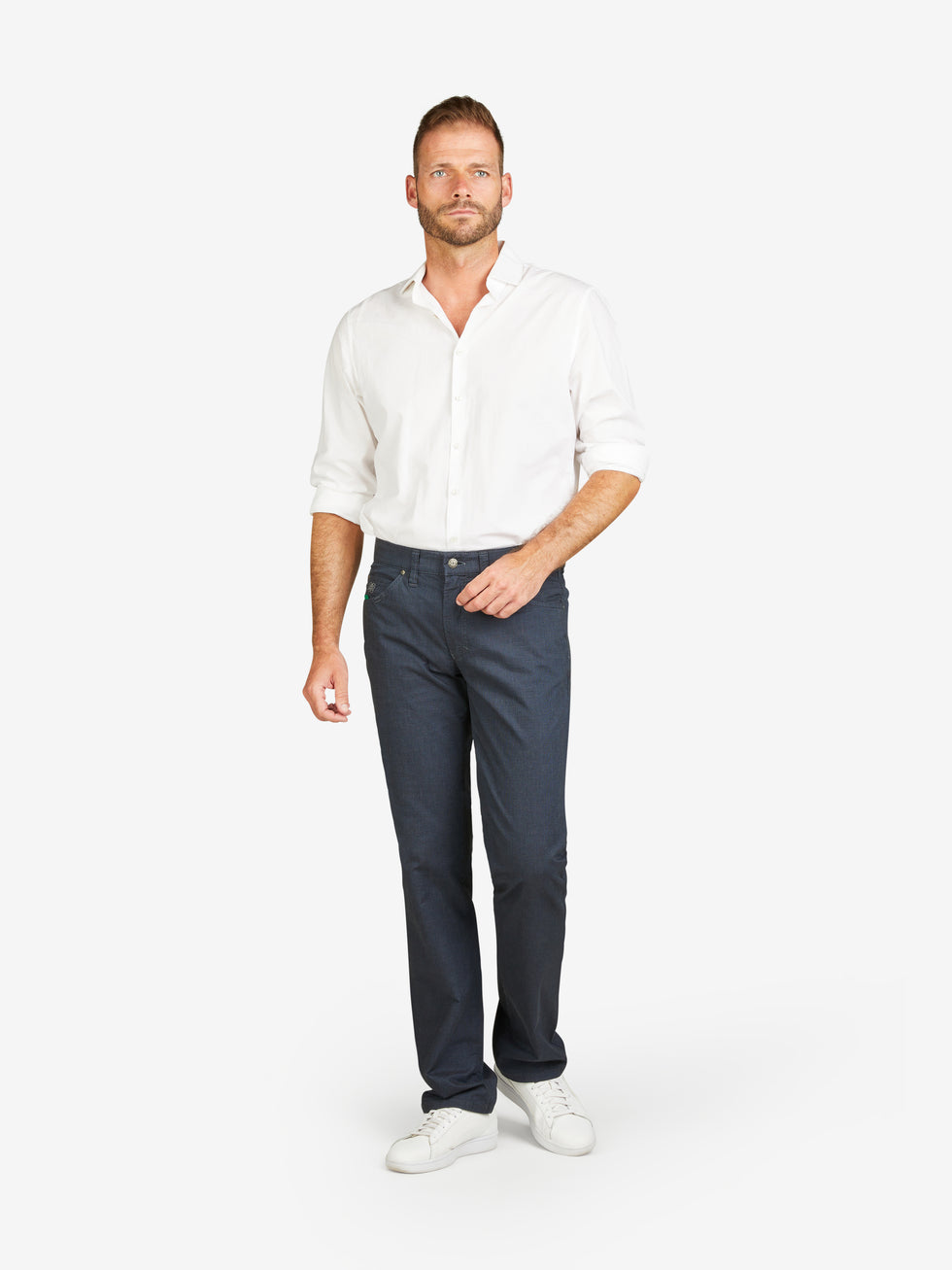 Mann trägt dunkelblaue HENRY 6910 5-Pocket-Hose aus Baumwoll-Mikrostruktur mit Wool-Look, elastischem Doppelstoffbund, Mobile Pocket in rechter Vordertasche, Kontrast-Münztasche, normaler Leibhöhe, schmalem Bein, weißes Hemd und Sneakers, neutraler Hintergrund.