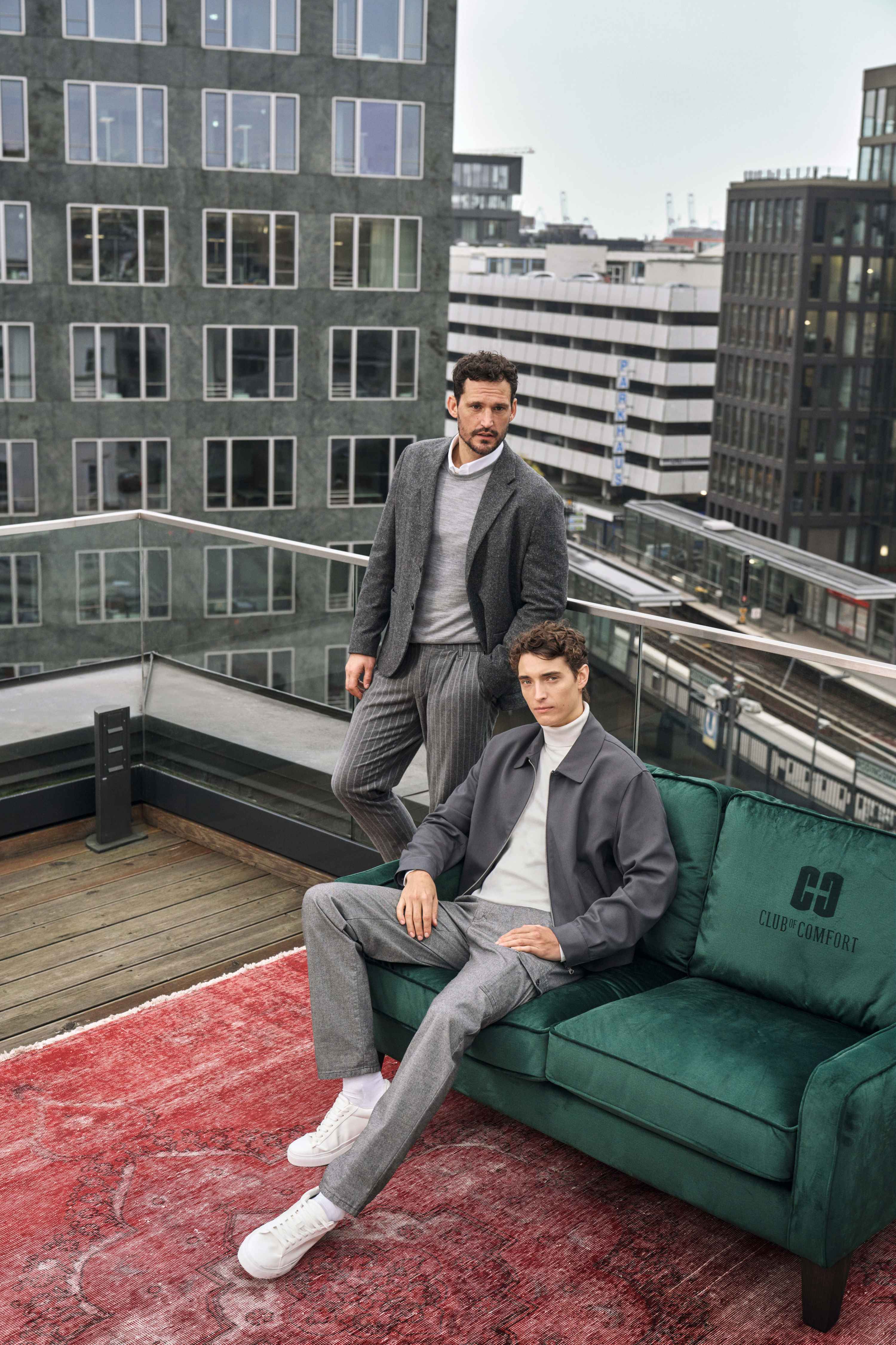 Zwei junge Männer mit dunklen, kurzen Haaren und durchschnittlicher Statur auf einer Dachterrasse in einer Stadt. Eine Person sitzt auf einem grünen Sofa, trägt einen grauen Anzug mit weißen Sneakers, die andere steht dahinter, trägt einen dunkelgrauen Mantel und eine helle Strickjacke. Im Hintergrund sind moderne Bürogebäude und ein Bahnsteig sichtbar.