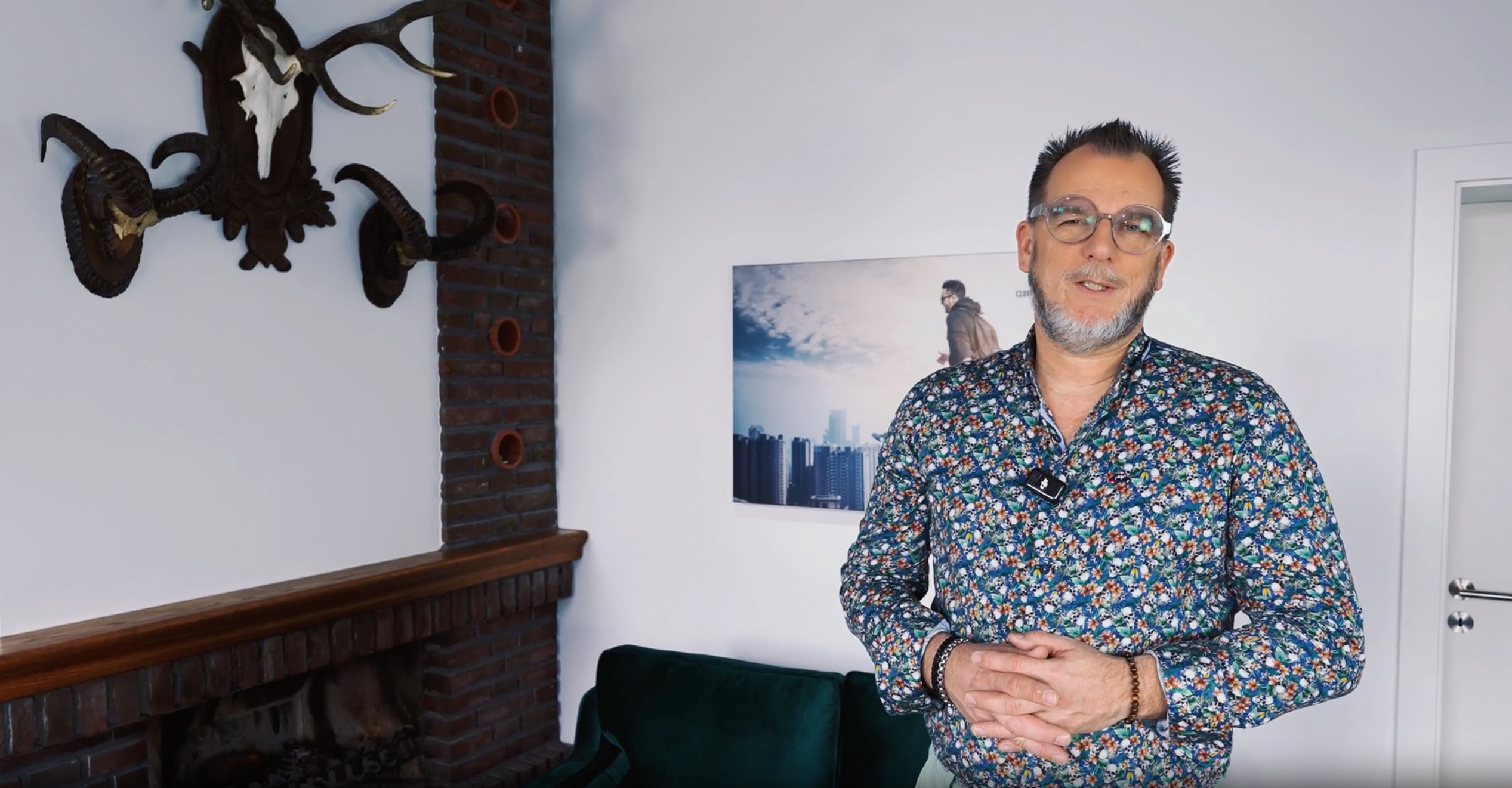Ein Mann mit Brille und Kurzhaarschnitt steht in einem modernen Wohnzimmer, trägt ein bunt gemustertes Hemd und lächelt. Im Hintergrund sind eine weiße Wand, ein Bild eines Stadtbilds, eine schwarze Giraffen-Schädeldekoration an der Wand und ein dunkelgrünes Sofa sichtbar.