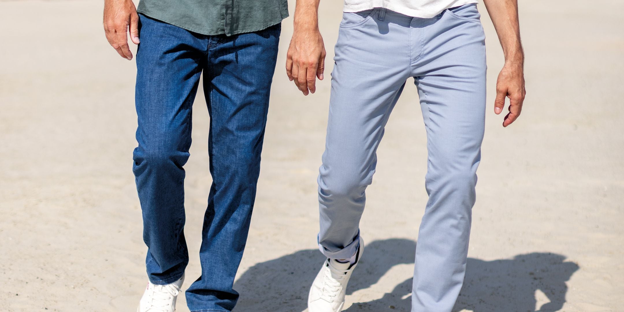Zwei Personen gehen Hand in Hand auf einer sandigen Oberfläche. Die Person auf der linken Seite trägt dunkle Jeans und einen grauen Oberteil, während die Person auf der rechten Seite helle Hellblaue Hosen und weiße Schuhe trägt.