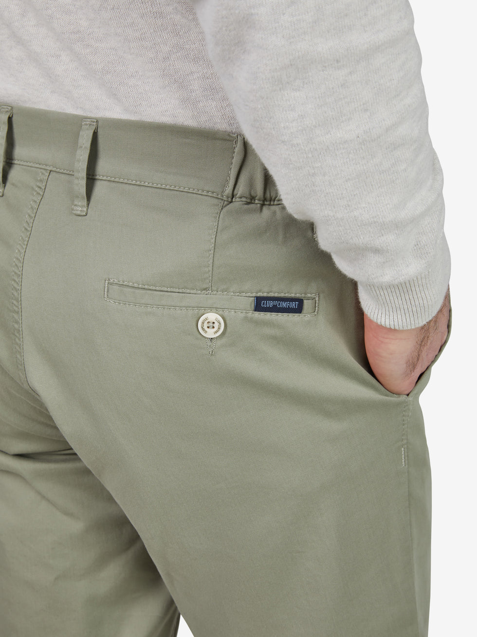 Nahaufnahme einer khakifarbenen Chinohose von Club of Comfort mit schmalem Bein, Comfort-Flex-Bund, Hemdenstopper, aus sommerleichter High Stretch Baumwolle mit 280 g/m², sichtbare Rücken-Reißverschlusstasche mit Label, getragen von einer Person in grauem Oberteil.