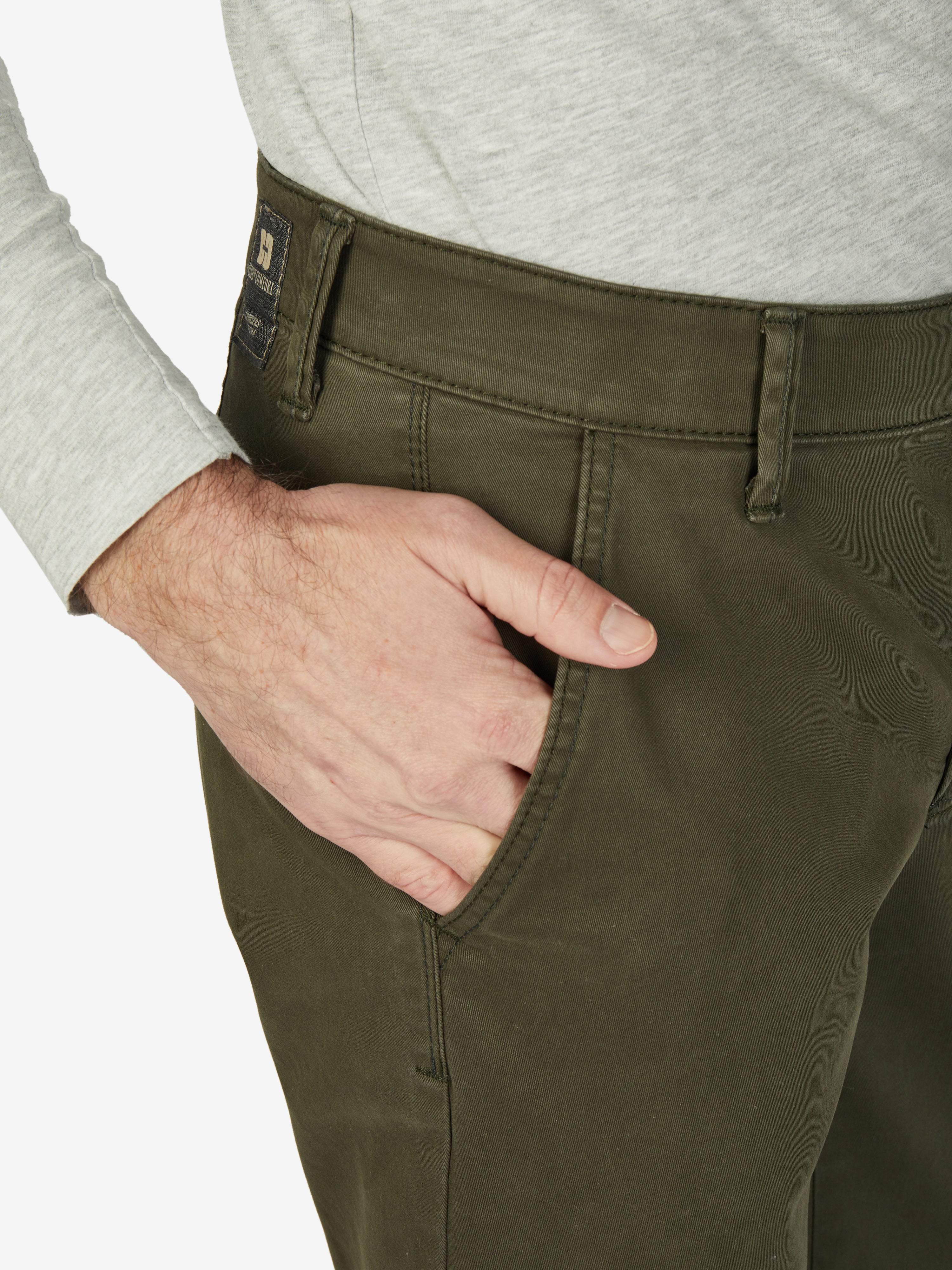 Nahaufnahme einer dunkel khakifarbenen Flat Front-Hose mit elastischem Komfortbund und Hemdenstopper, hergestellt von Club of Comfort. Die Hose hat eine normale Leibhöhe, schmalere Fußweite, ist aus hochelastischem Baumwoll-Gabardine mit High Stretch und wiegt 450 g/m. Ideale Chino für Herbst/Winter, mit Komfortbund, Made in Green by Oeko-Tex.