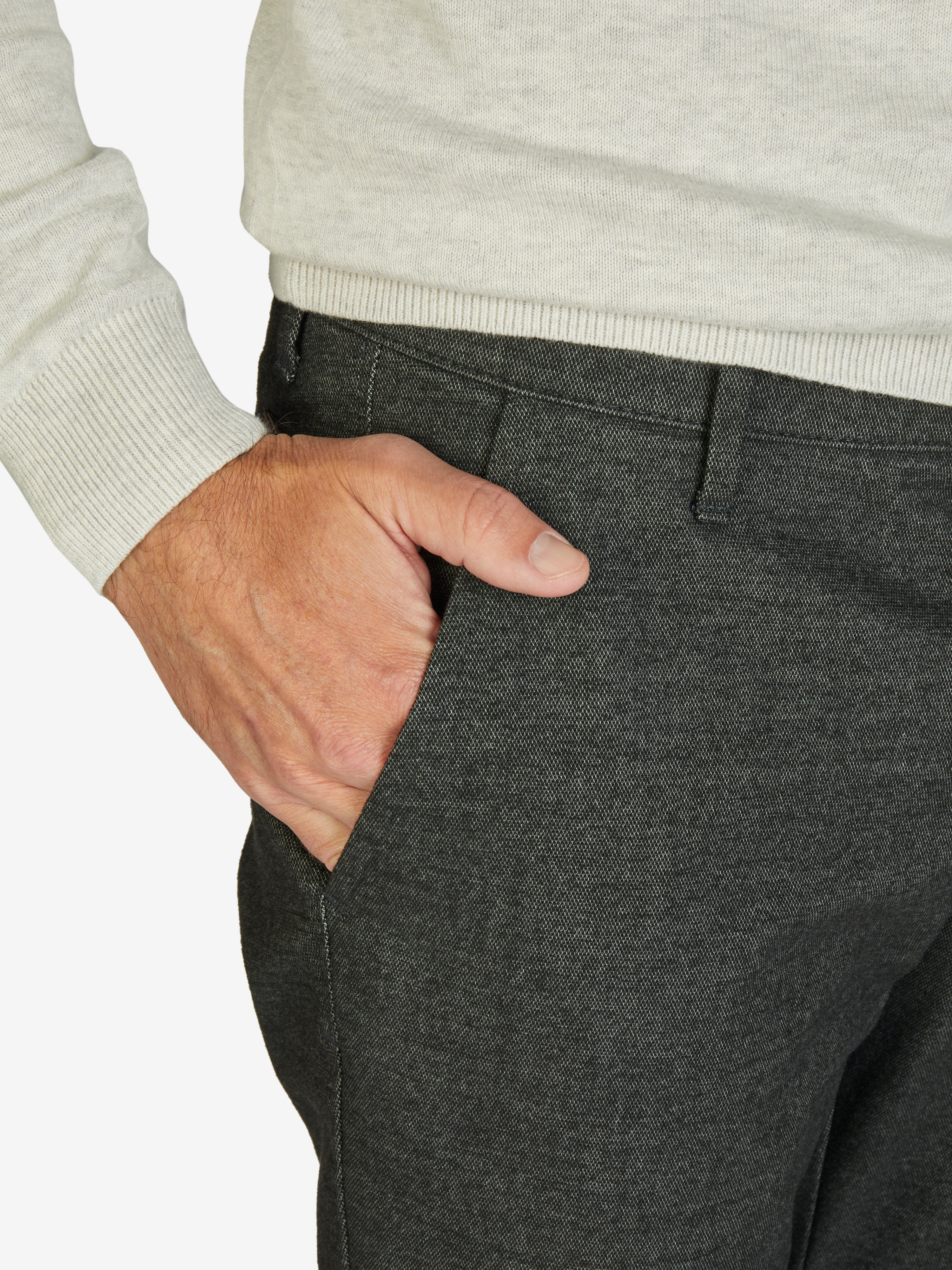 Nahaufnahme einer dunkelgrauen melierten Flat Front-Hose von GARVEY, mit elastischem Komfortbund und Hemdstopper, gefertigt aus Baumwoll-Struktur-Mischgewebe mit Woll-Optik, normaler Leibhöhe und schlanker Fußweite, getragen von einer Person in einem hellen Pullover.