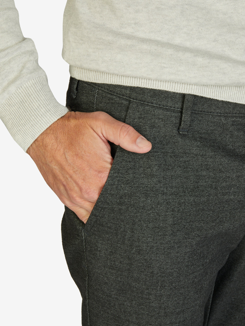 Nahaufnahme einer dunkelgrauen melierten Flat Front-Hose von GARVEY, mit elastischem Komfortbund und Hemdstopper, gefertigt aus Baumwoll-Struktur-Mischgewebe mit Woll-Optik, normaler Leibhöhe und schlanker Fußweite, getragen von einer Person in einem hellen Pullover.