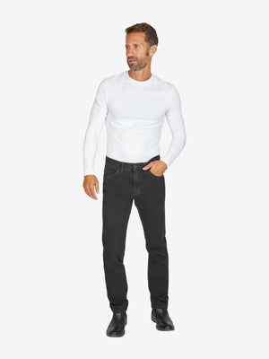 Mann trägt schwarze MARVIN 7054 Jeanshose von Club of Comfort in Regular Fit mit normaler Leibhöhe, elastischem Bi-Stretch-Denim, Swing-Pocket, Comfortbund, Hemdstopper, in Schwarz, model mit weißem Langarm-Shirt, schwarz-beige Schuhen, isoliert vor weißem Hintergrund.