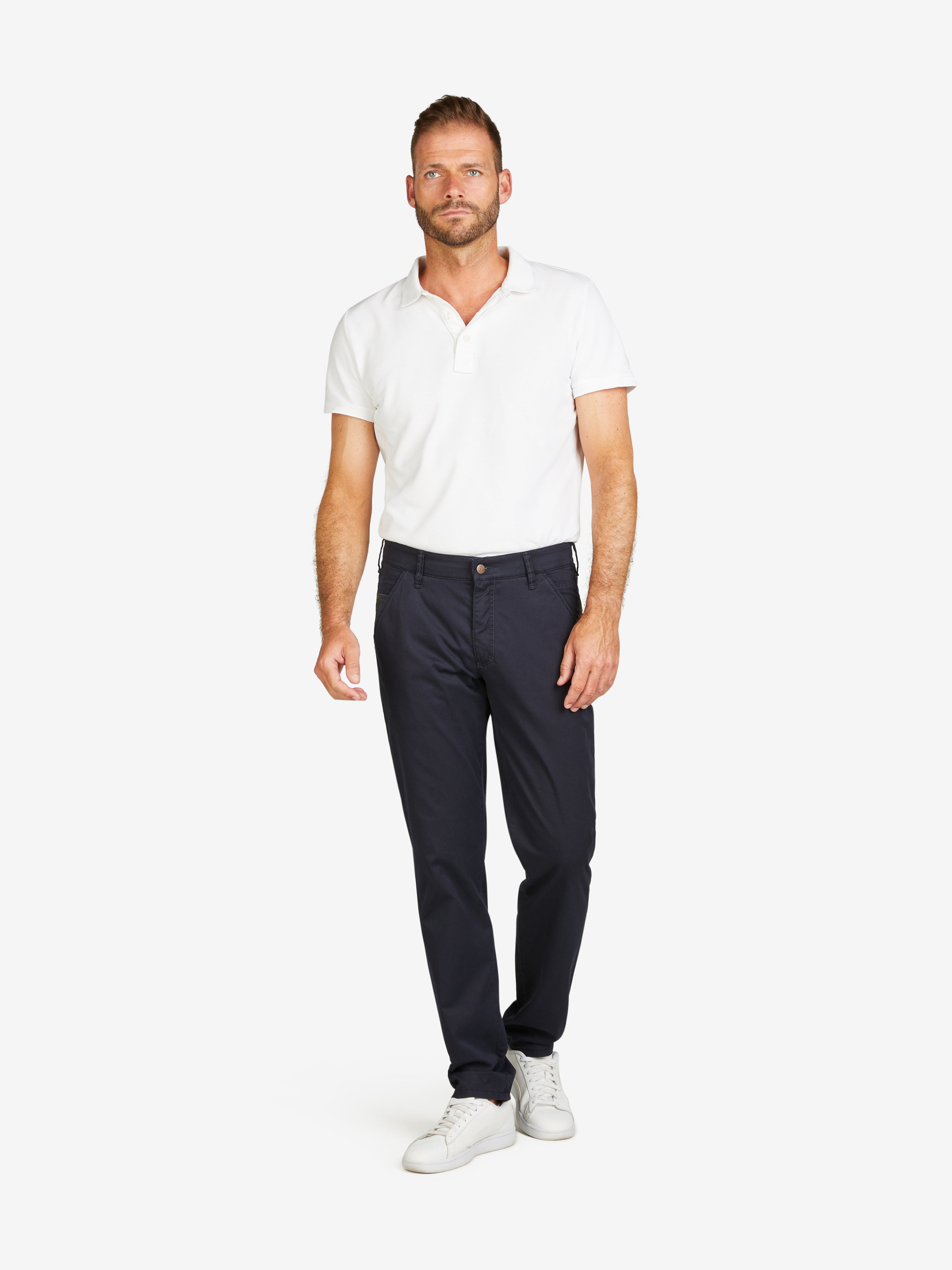 MännlicherModel trägt eine Marineblaue PHIL 7708 Chinohose von Club of Comfort mit normaler Leibhöhe, Slim Fit, elastischem Comfortbund, Hemdenstopper und Münztasche, kombiniert mit weißem Polohemd und weißen Sneakers, auf neutralem Hintergrund.