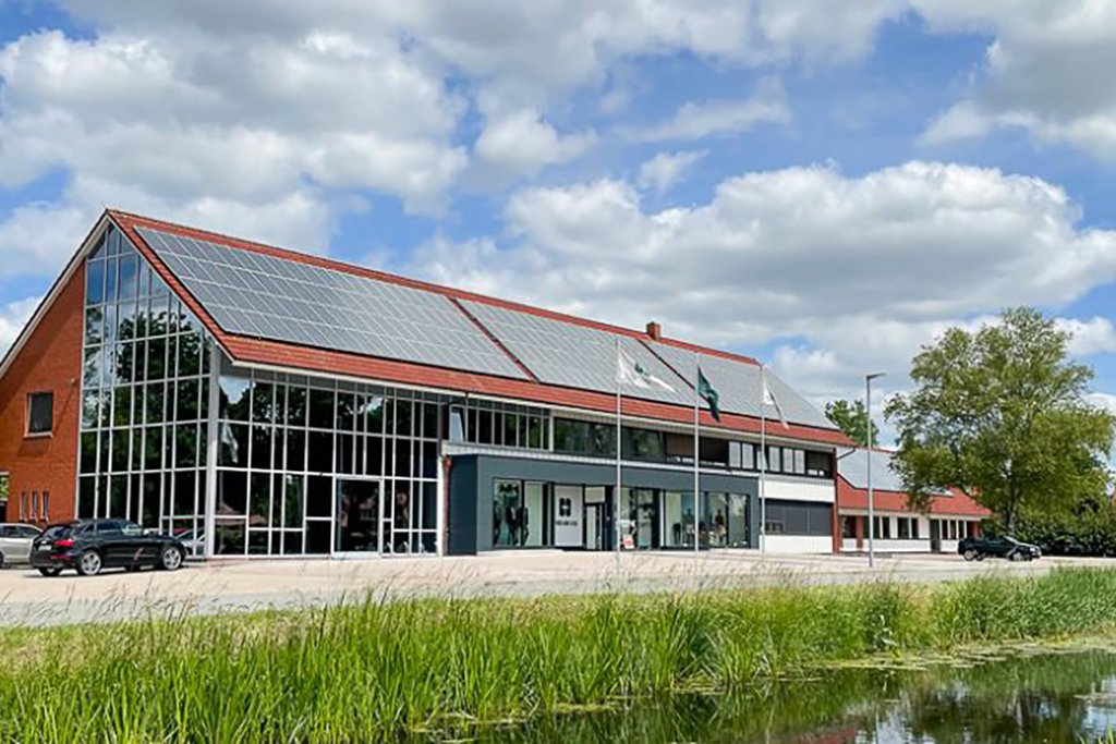 Das Bild zeigt ein modernes Gebäude mit einer steil geneigten Dachfläche, die vollständig mit Solarzellen bedeckt ist. Das Gebäude verfügt über große Fensterfronten und ist vor einem blauen Himmel mit einigen Wolken zu sehen. Im Vordergrund sind Gräser und ein Wasserlauf sichtbar, während einige Parkplätze mit Fahrzeugen vor dem Gebäude bestehen.