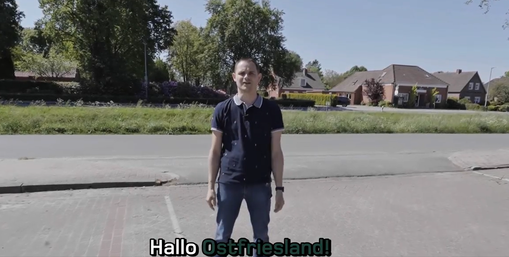 Ein junger Mann steht auf einem Gehweg neben einer Straße in einer ländlichen Gegend. Im Hintergrund sind Bäume, Einfamilienhäuser und eine Wiese zu sehen. Er trägt ein dunkelblaues T-Shirt und jeansblaue Hosen. Die Bildunterschrift lautet: "Hallo Ostfriesland!".