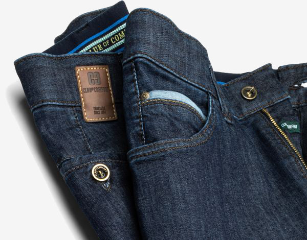 Nahaufnahme einer dunkelblauen Jeans mit sichtbaren Nähten, Metalldruckknöpfen und einem kleinen Reißverschluss in der Tasche. Das Innenetikett zeigt das Markenlogo "G-STAR RAW" und der Gürtel ist teilweise sichtbar.