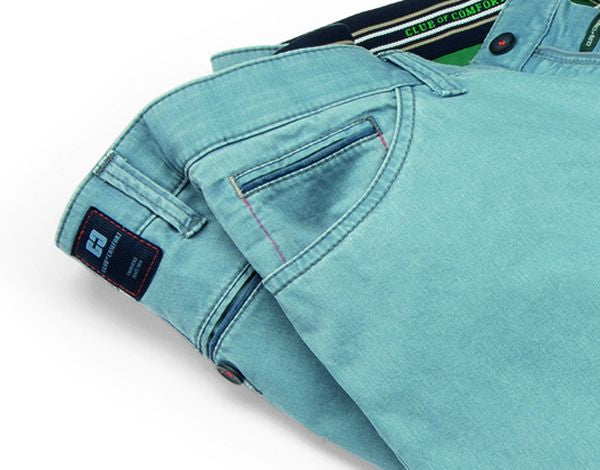 Hellblaue Denim-Hose mit sichtbaren Nähnähten in mehreren Farben, eine waagerechte Brusttasche mit Reißverschluss, einem kleinen Knopf an der Taille, einem grünen Label im Bund und einem dunklen Etikett an der Rückseite.