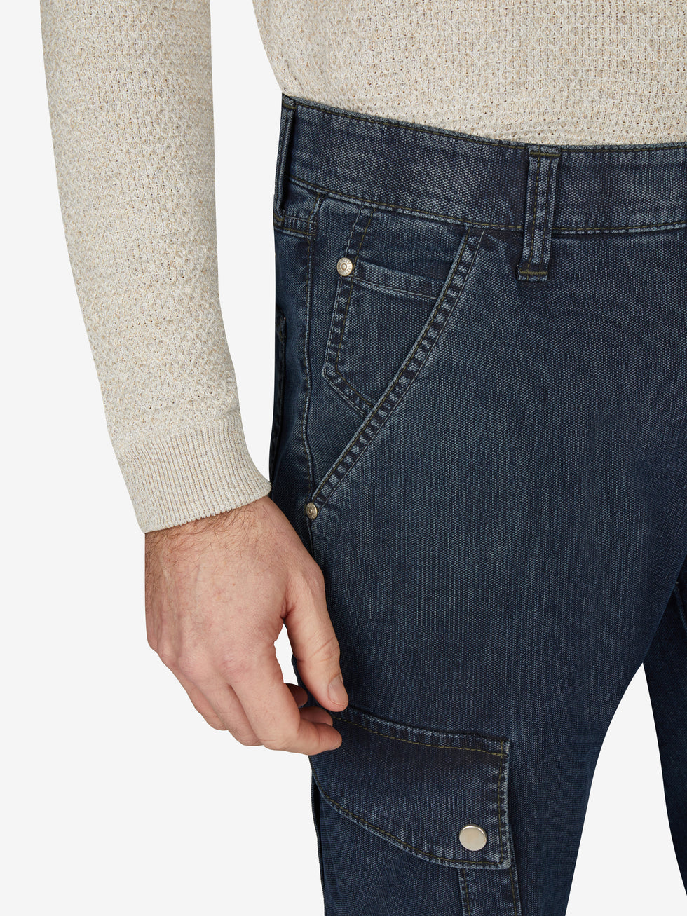 Detailliertes Produktbild einer blauen TANGO 8012 Cargohose im Tapered Fit mit elastischem Comfortbund, Hemdenstopper, aufgesetzten Gesäßtaschen, Beintaschen mit Patte und Druckknopf, aus High Stretch Denim mit filigranem Muster, gewichtet 460 g/m, hoher Tragekomfort, Made in Green by Oeko-Tex, für Herbst/Winter, sichtbar an einer Person im beige Pullover.
