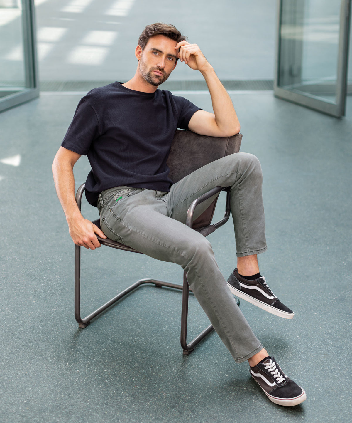 Ein junger Mann mit dunklem Haar und Bart sitzt entspannt auf einem Stuhl in einem modernen, hell erleuchteten Raum mit großen Fenstern. Er trägt ein schwarzes T-Shirt, graue Jeans und schwarze Vans-Sneaker, wirkt nachdenklich und relaxed.