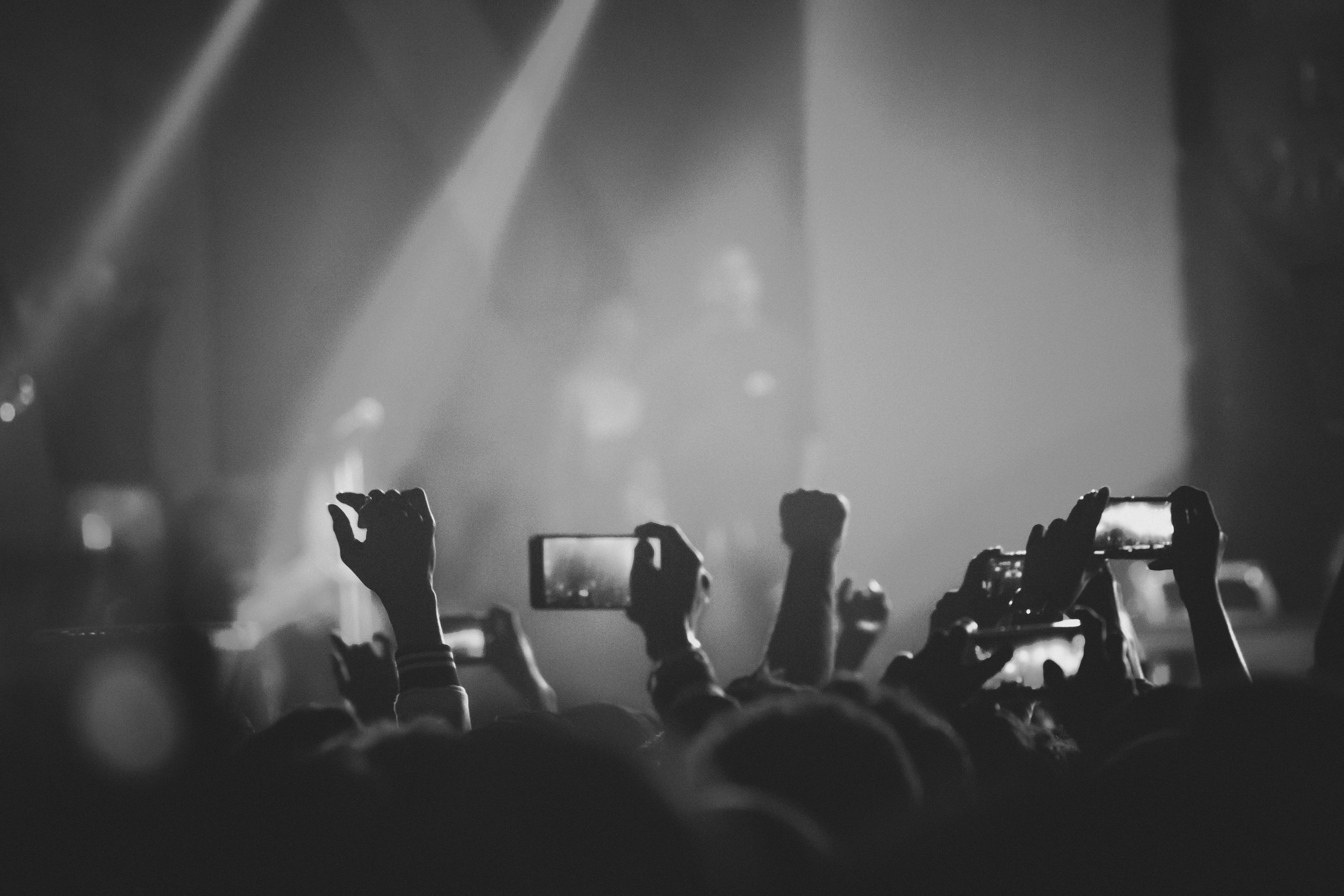 Menschen im Publikum eines Konzerts halten ihre Smartphones hoch, um Fotos oder Videos von der Bühne aufzunehmen. Im Hintergrund sind Scheinwerfer und eine dunkle Konzertatmosphäre sichtbar.